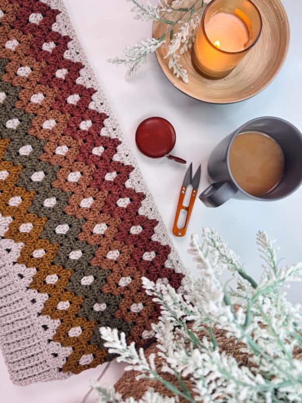 A multi-colored crochet blanket, small red container, scissors, gray mug with a beverage, candle in a wooden holder, and faux foliage on a white surface.