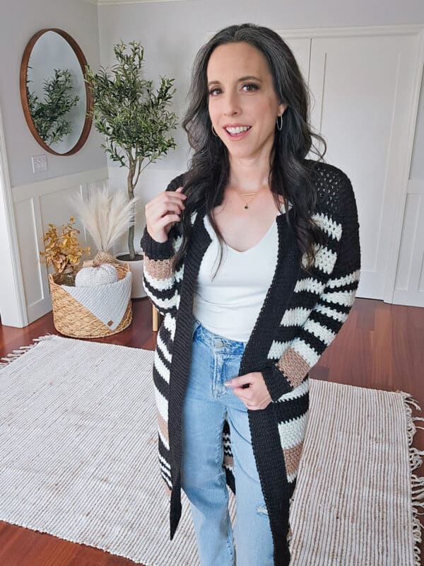 A woman with long hair stands indoors on a woven rug, wearing a white top, striped cardigan, and blue jeans. A round mirror, wall decor, and a plant are visible in the background. Her shortbread crochet duster adds a touch of elegance to the cozy setting.