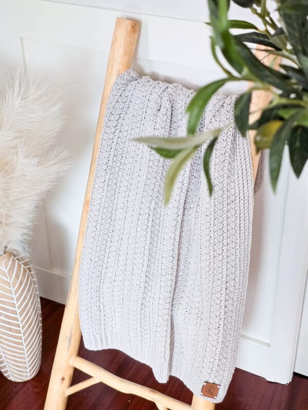 A braided wheat crochet blanket draped over a wooden ladder with plants nearby.