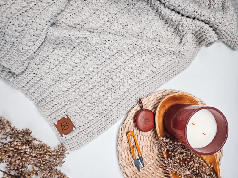 A cozy gray knit blanket, a lit candle in a red holder on a wicker coaster, dried flowers, and small tools are arranged on a white surface.