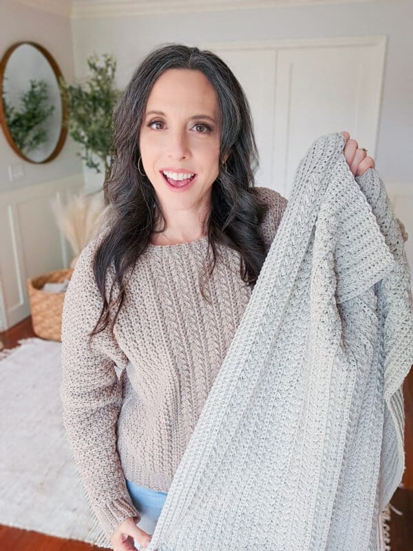 A person with long dark hair and a gray knitted sweater holds up a matching knitted blanket. They are smiling and standing in a well-lit room with a mirror and basket in the background.