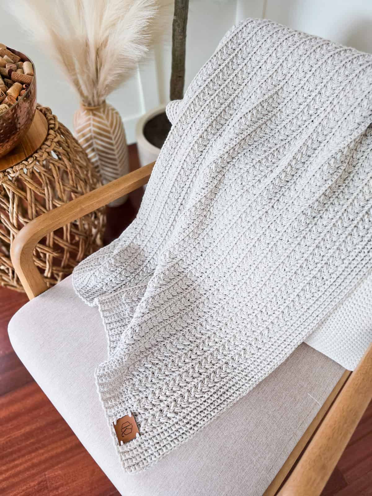 A knitted gray blanket draped over a wooden chair with a light beige cushion, next to a woven basket of pinecones and dried pampas grass adornments on a wooden floor.