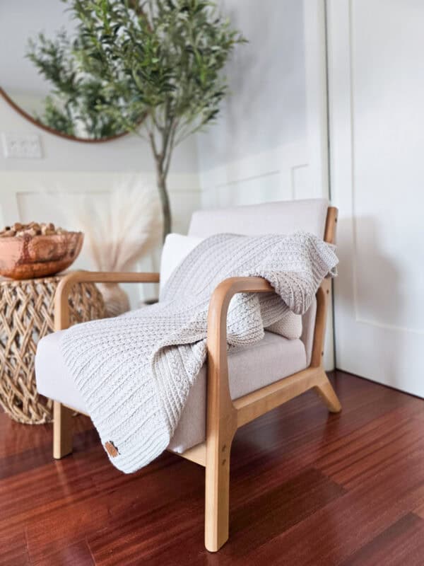 A wooden armchair with a white knitted blanket sits on a polished wooden floor next to a round wicker table and a potted plant in the background.