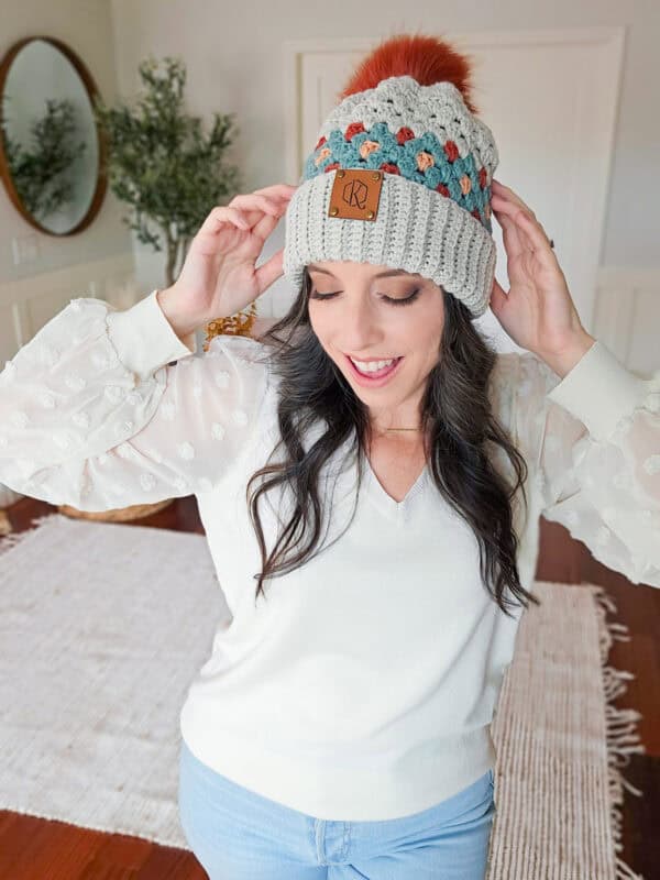 A woman in a white blouse and jeans stands indoors, adjusting her multicolored knit beanie. She smiles softly with closed eyes.