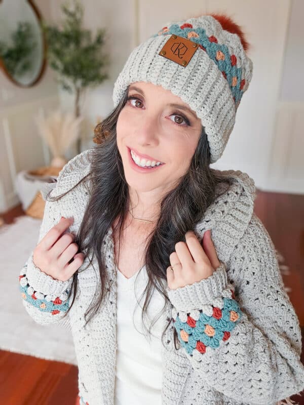 A woman wearing a gray knit hat and matching cardigan with colorful accents smiles indoors.