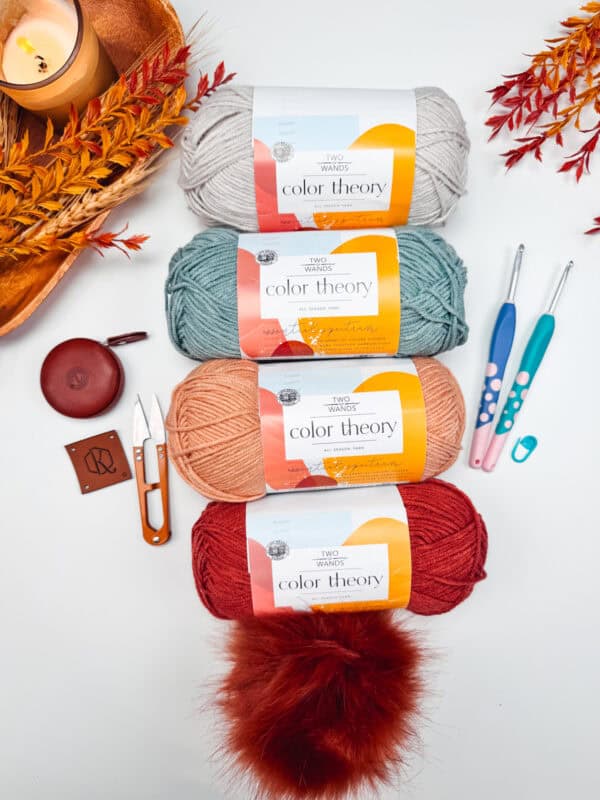 Four skeins of "Color Theory" yarn in various colors are arranged vertically alongside crochet hooks, scissors, a measuring tape, and decorative leaves on a white surface.