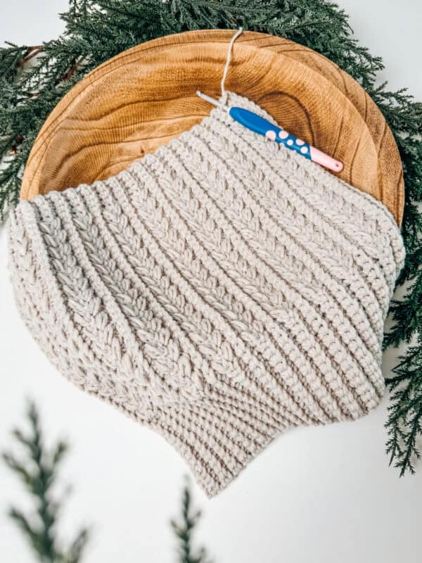 Knitted cream shawl in progress with a crochet hook lies on a wooden plate, surrounded by evergreen branches.