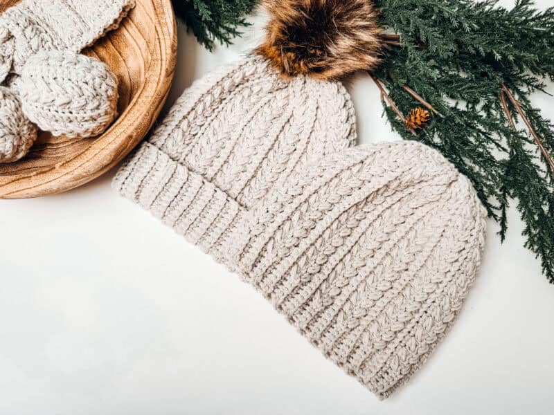 Two knitted cream-colored beanies with faux fur pom-poms rest on a white surface, surrounded by green pine branches and a wooden bowl of yarn. These cozy accessories evoke the charm of a snowy day, akin to creating your own Wheat Puff crochet hat pattern.