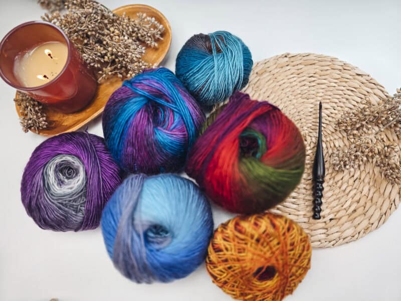 Colorful yarn balls on a table with dried flowers, a lit candle, a woven mat, and a black crochet hook.