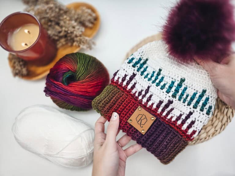 A person holds a colorful knitted hat with a pom-pom, surrounded by yarn and a lit candle on the table. The intricate design resembles the Mosaic Ridge Crochet Hat Pattern, adding an artistic touch to the cozy scene.