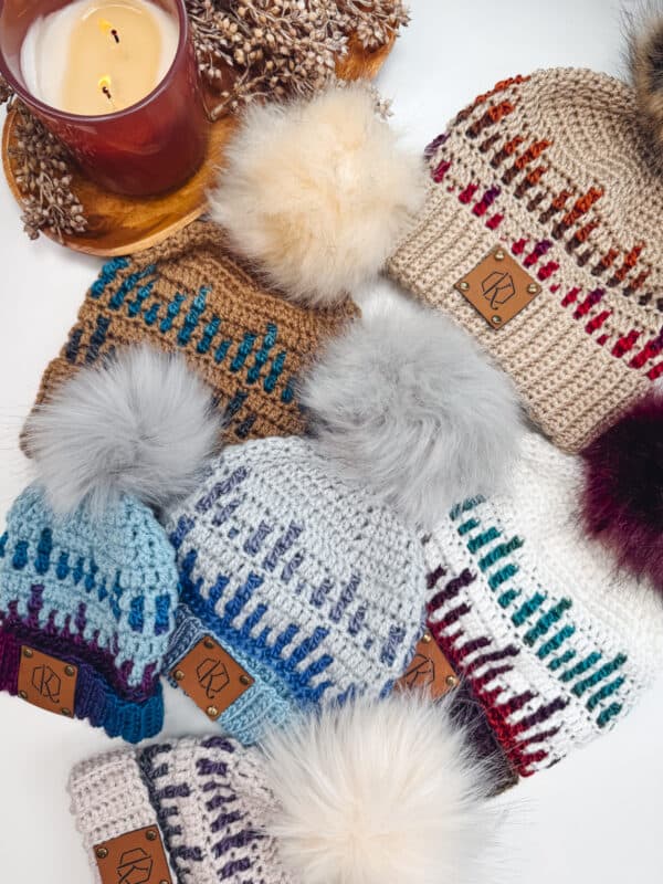 A collection of knitted hats with pom-poms, featuring various multicolored patterns, arranged on a surface next to a lit candle.