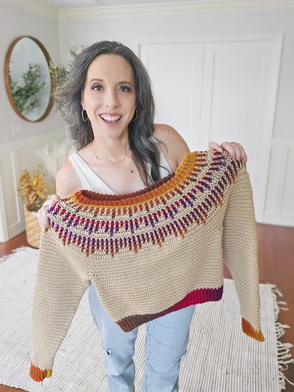 A person holds up a knitted sweater with a colorful patterned yoke, standing in a room with wooden floors and a mirror on the wall.