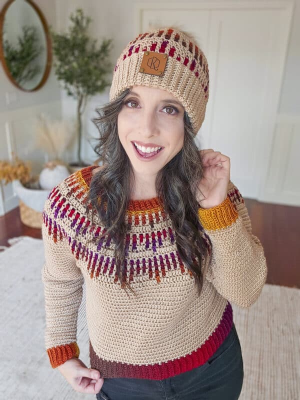 A person wearing a knitted sweater and matching beanie with red and orange patterns, standing indoors and smiling.