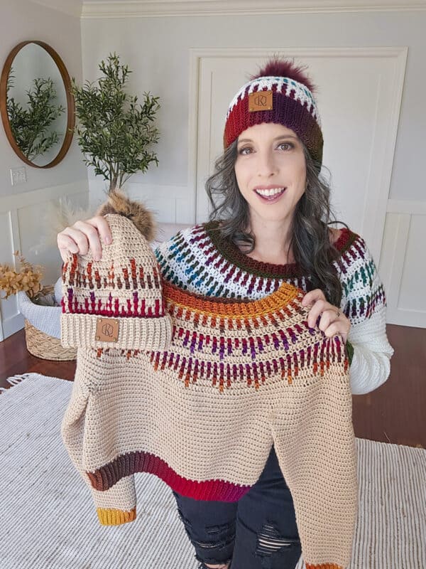 Woman smiling, holding a knitted sweater and matching hat with colorful patterns. Another hat rests on her other hand. Room has a mirror, plant, and light decor.