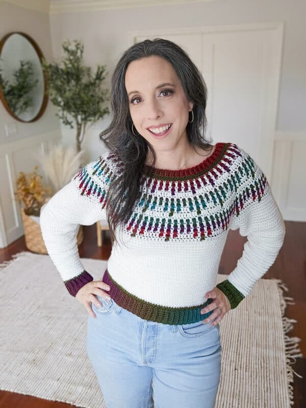 A person wearing a colorful knitted sweater and jeans stands in a room with wooden floors and a rug, smiling at the camera.