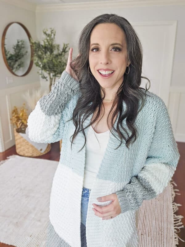 A woman with long dark hair in a white and blue color block crochet duster stands in a room with a round mirror and a potted plant.