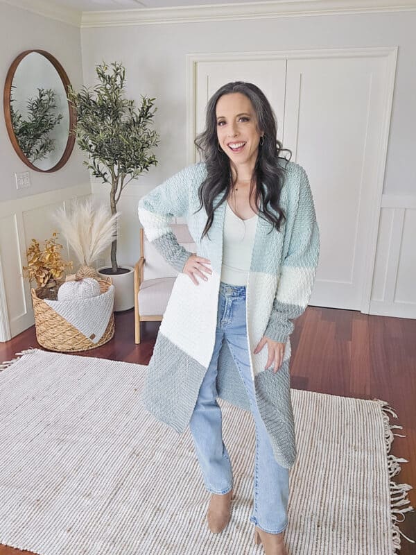 A person stands in a room with a plant, wearing a color block crochet duster over light jeans and boots, smiling at the camera.