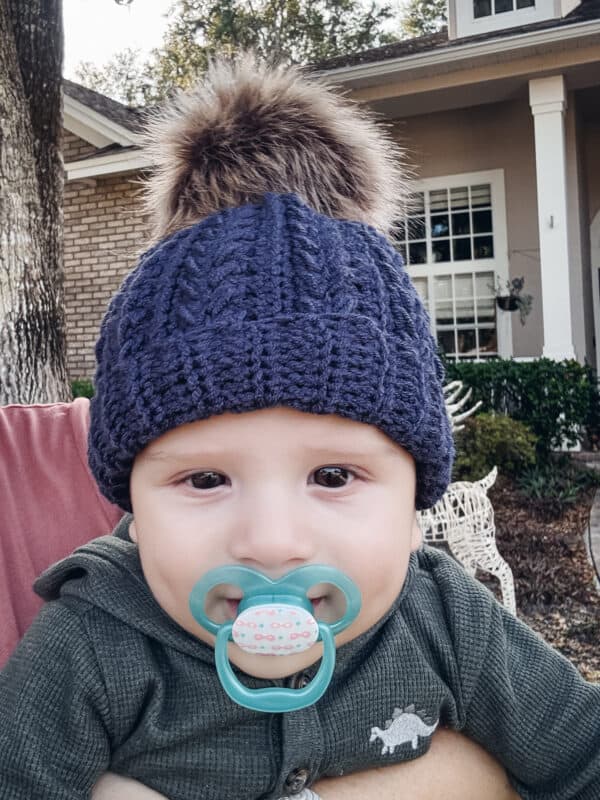 A baby wearing a knitted hat with a fur pom-pom and a pacifier in their mouth, sitting outside in front of a house.