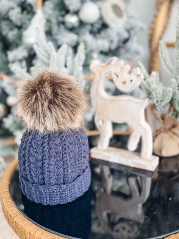 A knitted hat with a faux fur pom-pom sits on a table. A wooden reindeer decoration and a frosted Christmas tree are in the background.