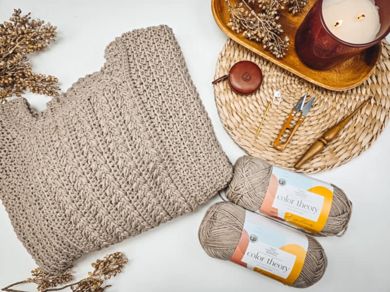 A knitted beige sweater is accompanied by two skeins of yarn, knitting tools, dried flowers, a lit candle, and a woven mat arranged neatly on a white surface.