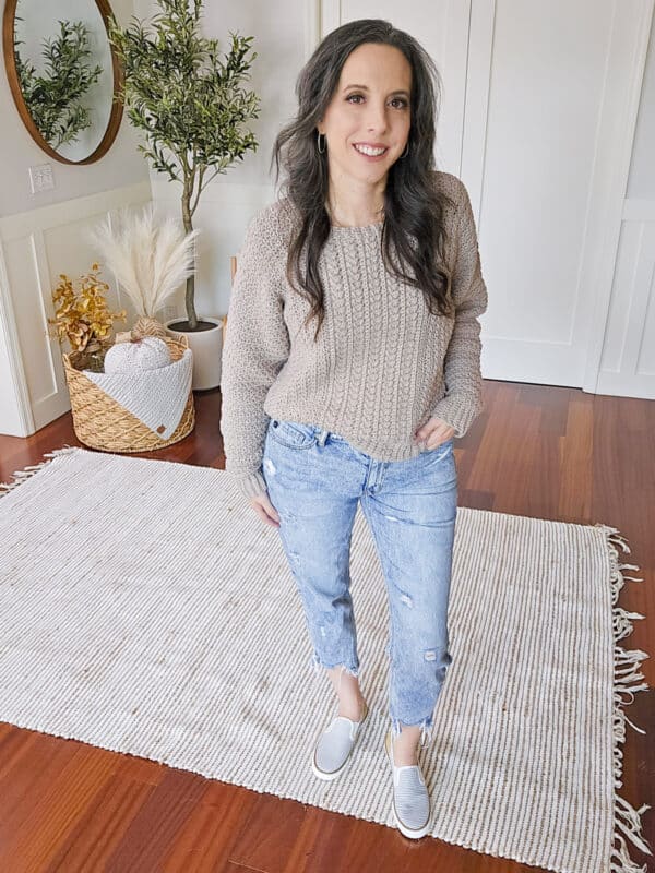 A woman with long dark hair wearing a beige knit sweater, light blue distressed jeans, and gray shoes stands on a white rug in a room with wooden floors and a plant basket.