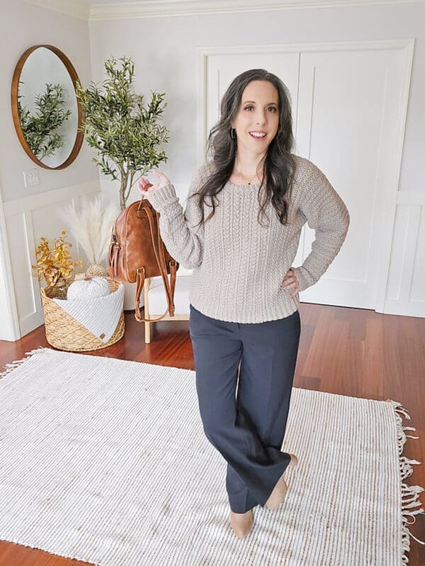 A woman with long dark hair stands in a room with wooden floors, wearing a light gray sweater and dark pants. She is smiling and gesturing with her left hand. There is a round mirror and potted plant behind her.