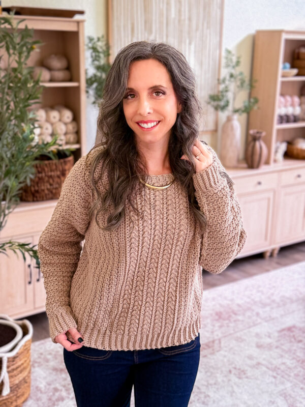 A woman with long wavy hair wearing a toasty wheat crochet sweater and a gold collar necklace.
