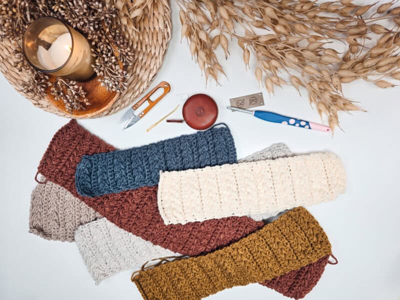 Knitted scarves in various colors are laid out on a table with crochet tools, a measuring tape, a candle, and dried foliage.