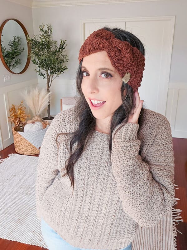 A person with long dark hair, wearing a beige sweater and a brown knitted headband, poses indoors in front of a white wall and a mirror.