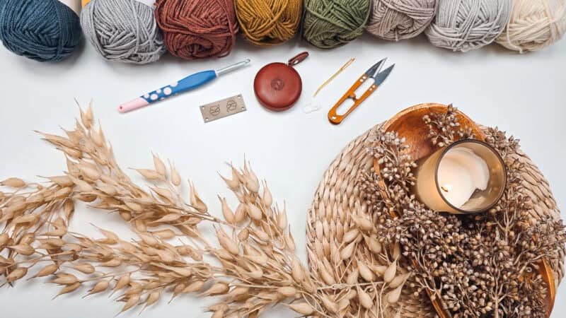 Colored yarn, crochet tools, scissors, measuring tape, a metal needle gauge, a lit candle, and dried plants arranged on a white surface.