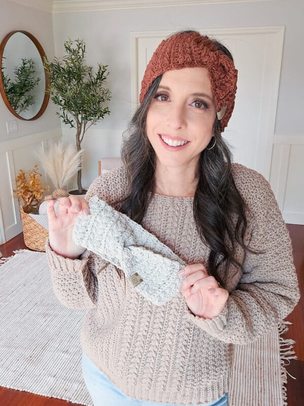 A woman in a cozy sweater and brown headband smiles while holding a gray knitted headband. She stands in a room with a round mirror and indoor plants.