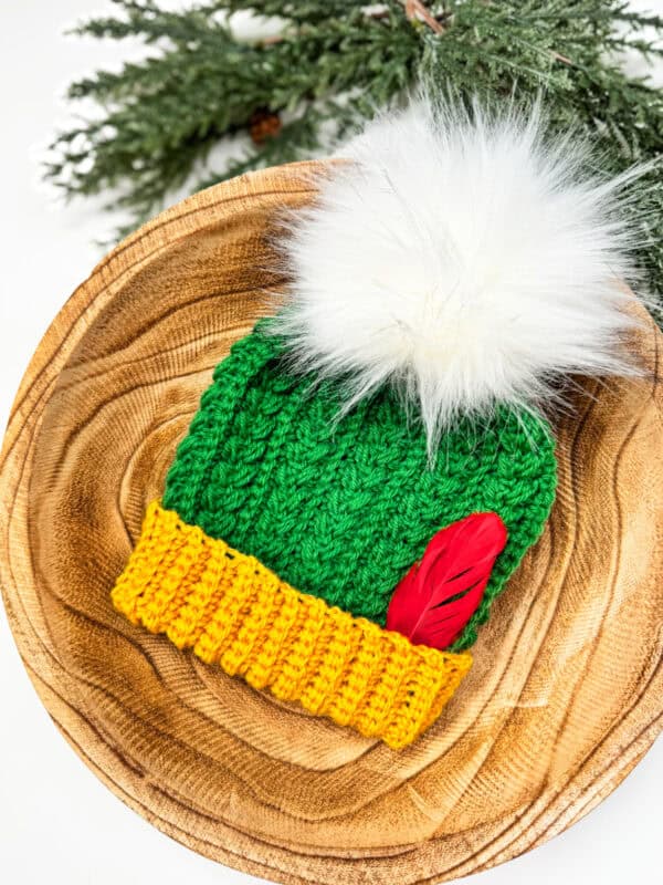 A snowy green knitted hat with a yellow brim, white pom-pom, and red feather rests on a wooden plate. Pine branches frame the scene, evoking the charm of a Wheat Puff crochet pattern.