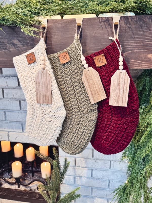 Three knitted holiday stockings (cream, green, and red) hang from a mantel above a fireplace with lit candles and garland.