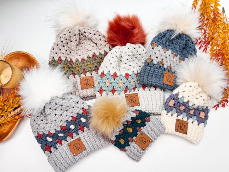 A variety of crocheted hats in gemstone hues, complete with pom-poms, are displayed surrounded by autumn leaves and a candle. Perfect the cozy vibe with a free pattern to craft your own unique beanie.