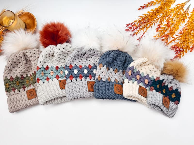 Five crocheted beanies in vibrant gemstone hues with colorful patterns and faux fur pom-poms are lined up. Autumn leaves and a candle set the cozy background, offering inspiration for your own creations. Explore our free pattern to craft these stunning crochet beauties yourself.