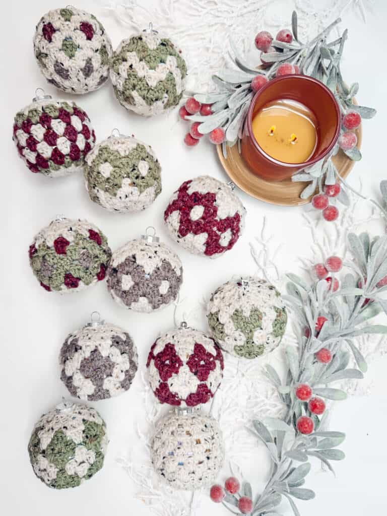 Crocheted ornaments in green, red, and white patterns are arranged beside a lit candle on a saucer, surrounded by frosted foliage with red berries.