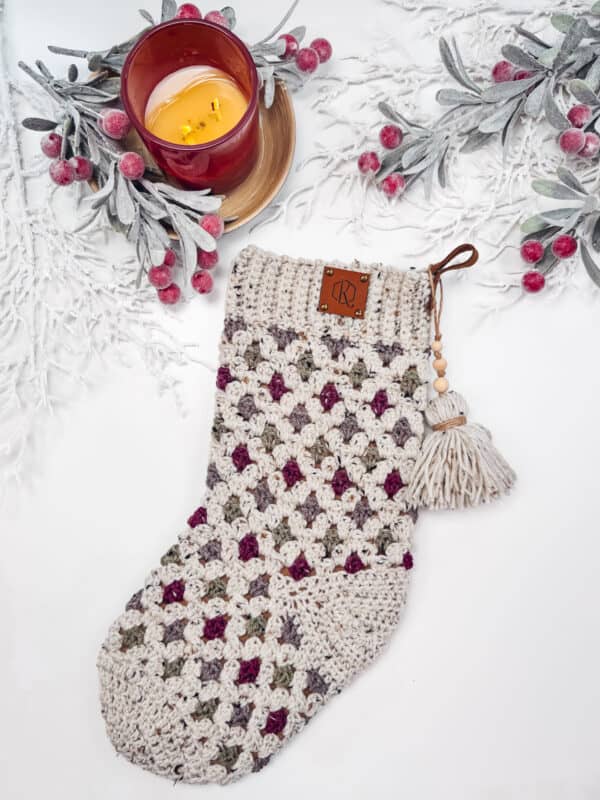 Crocheted stocking with tassels, featuring a multicolored pattern in beige, purple, and green, next to a lit candle in a red holder, surrounded by frosted leaves and berries.