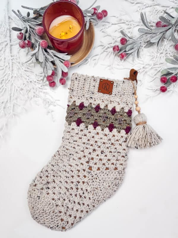 A crocheted Christmas stocking with green and purple accents, tassel, and leather tag. It's placed on a white surface with a red candle and frosted berry branches.