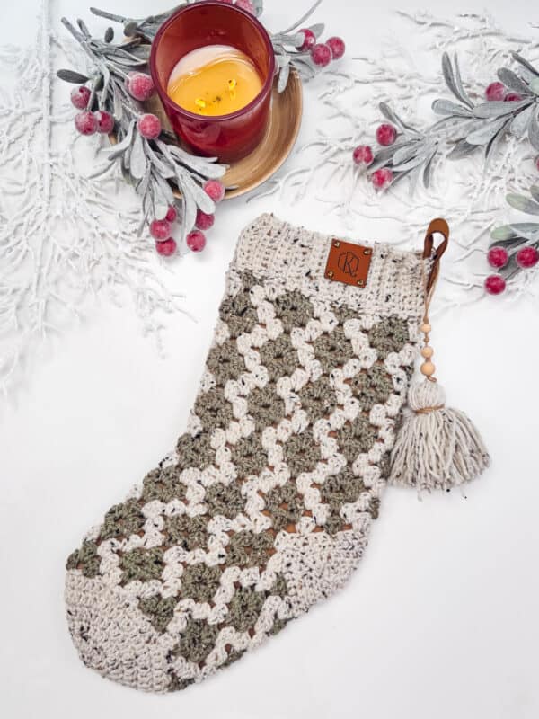 Crocheted stocking with tassel, featuring a rustic pattern, placed on a white surface adorned with frosted branches and red berries. A lit red candle in a glass holder is beside it.