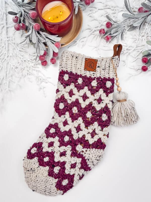 Knitted Christmas stocking with a white and burgundy pattern, a tassel, and a candle surrounded by frosted greenery and red berries.