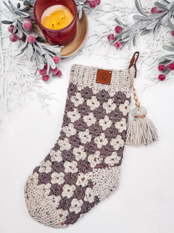 A crocheted Christmas stocking with a checkered pattern in gray and white, adorned with a tassel, lies on a white surface. A lit candle and frosted branches with berries are nearby.