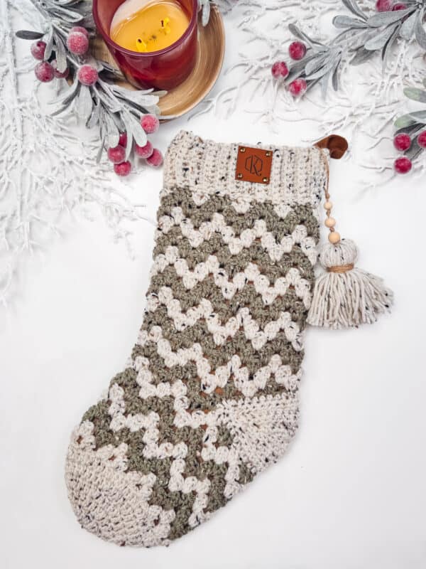 A knitted stocking with a zigzag pattern lies on a white surface, surrounded by frosted branches and berries. A lit candle in a red holder is nearby.