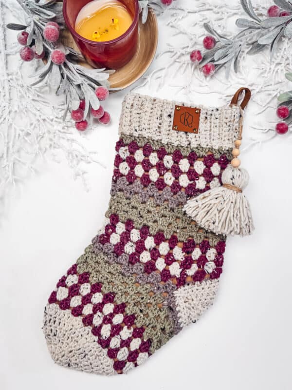 A crochet Christmas stocking with a tassel sits next to a lit candle and frosted branches with red berries.