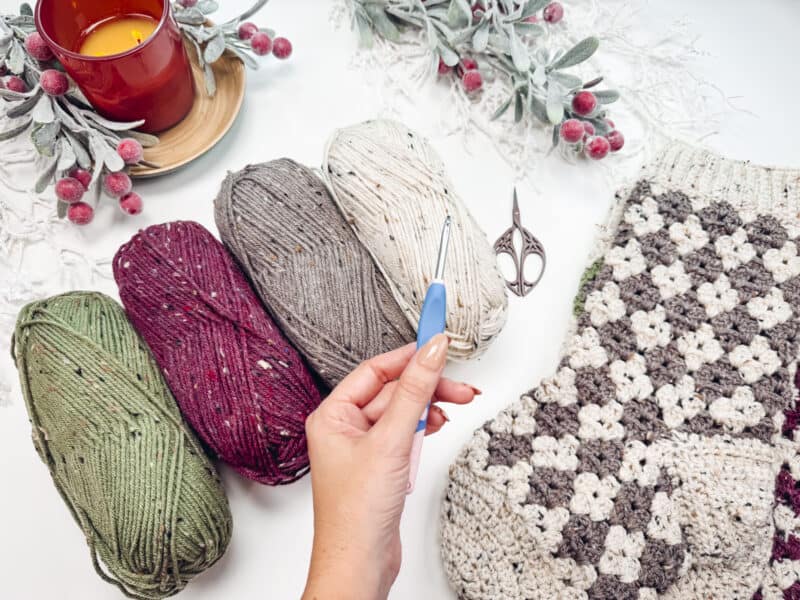 A hand holds a crochet hook above colorful yarn balls and a partly crocheted piece. A red candle and decorative berries adorn the white surface.