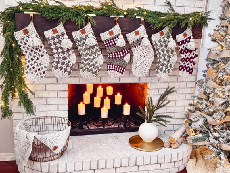 Cozy fireplace with lit candles, decorated with festive stockings and greenery. A lit Christmas tree and logs are nearby, with a basket holding a blanket on the hearth.