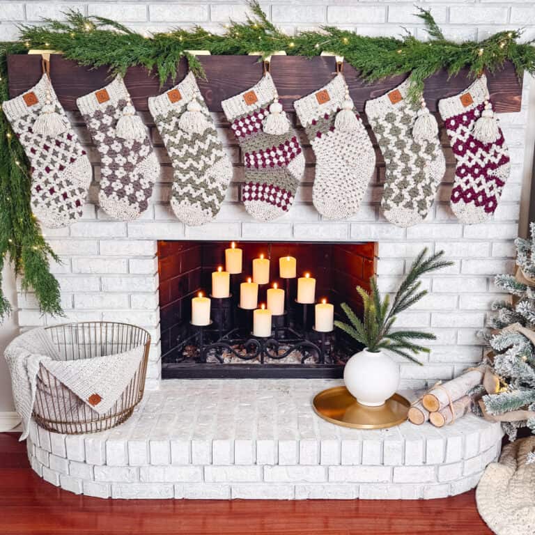 A white brick fireplace adorned with six stockings from the Granny All the Way Crochet Stocking Collection adds charm above lit candles inside. On the hearth, a basket with a blanket and a vase of greenery complete this cozy scene.