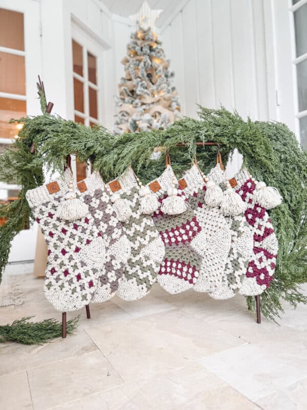 Five knitted stockings hanging on a rack decorated with greenery, with a decorated Christmas tree visible in the background.