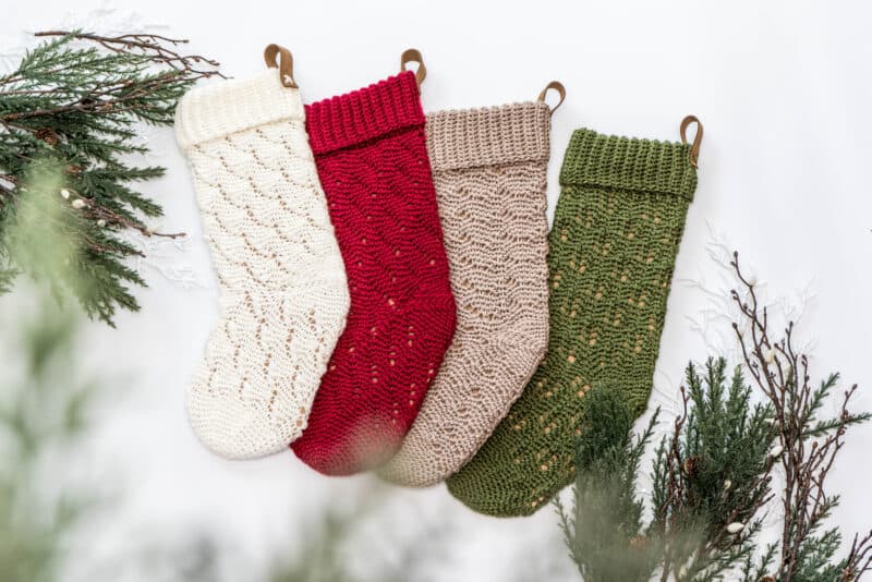 Four knitted Christmas stockings in cream, red, beige, and green hang against a white background, surrounded by greenery.