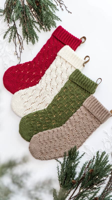 Four crocheted Christmas stockings in red, white, green, and beige, arranged on a white background with pine branches.