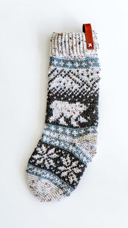 Knitted Christmas stocking with a polar bear design, featuring shades of gray, blue, and white, and a brown loop at the top.